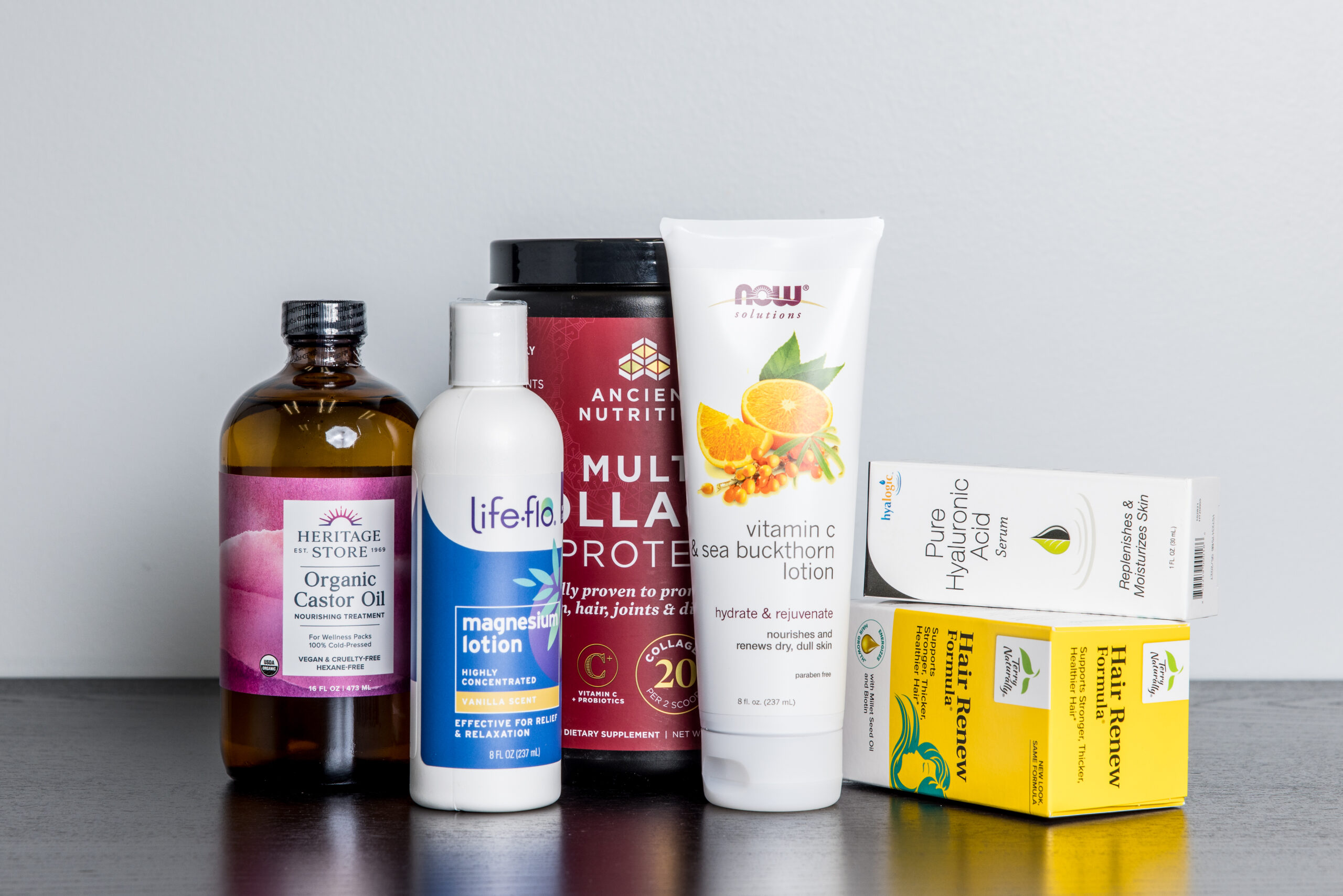 Six health and wellness products, including castor oil, magnesium lotion, collagen protein powder, vitamin C lotion, hyaluronic acid, and vitamin D3 and K2 supplements, are arranged on a dark surface against a white background.