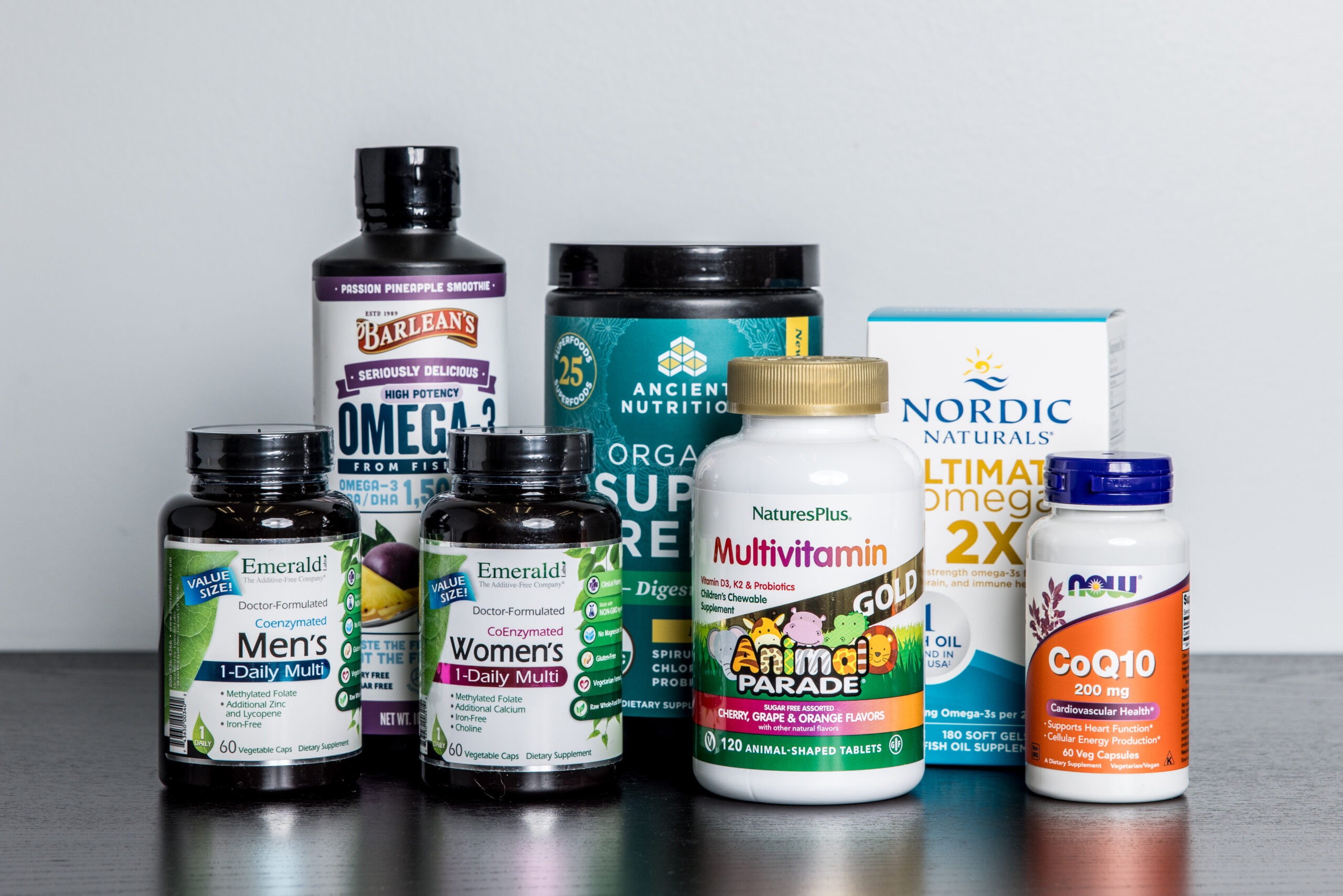 A variety of supplement bottles, including multivitamins, omega-3, CoQ10, and super greens, are arranged on a white surface against a gray background.