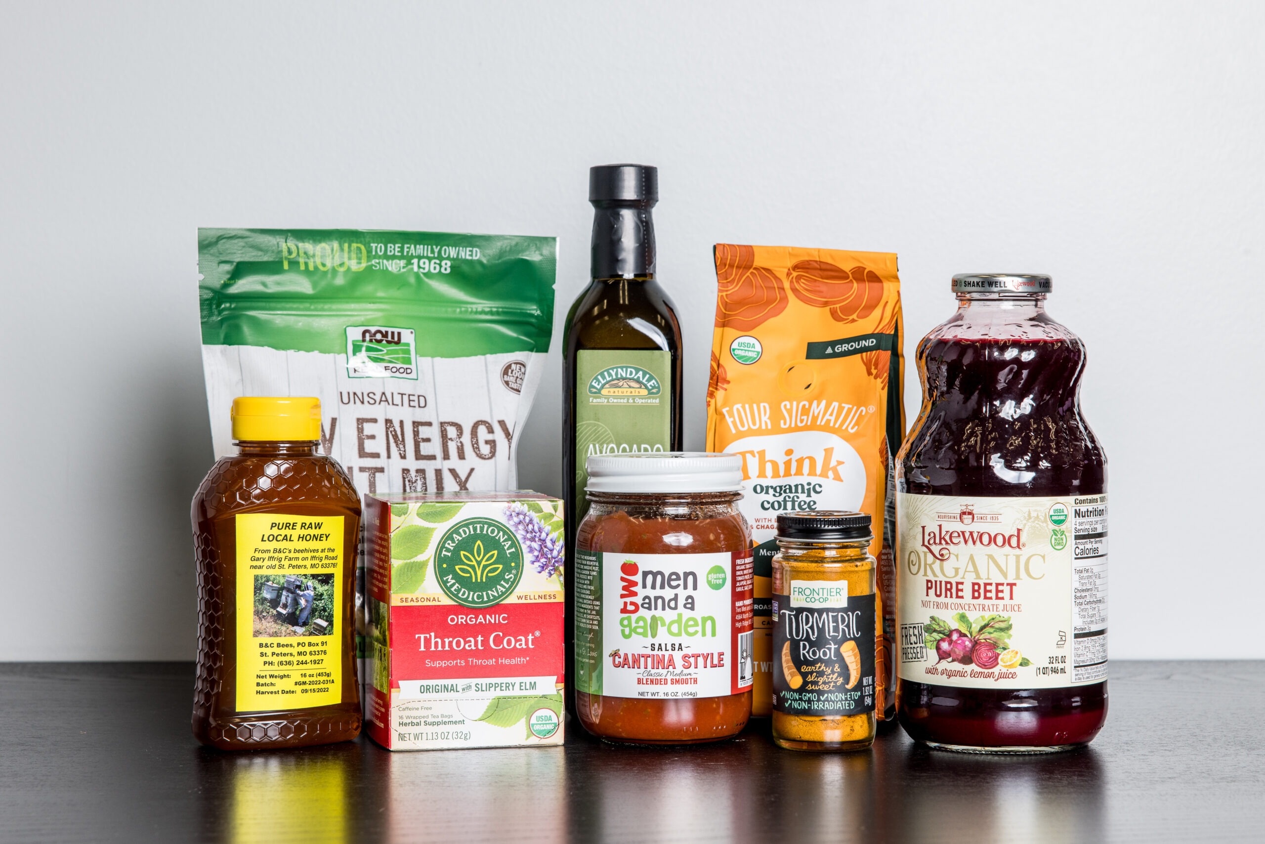 A variety of organic and health food products, including tea, olive oil, honey, energy mix, coffee, pasta sauce, turmeric root, and beet juice, arranged on a dark surface against a plain light background.